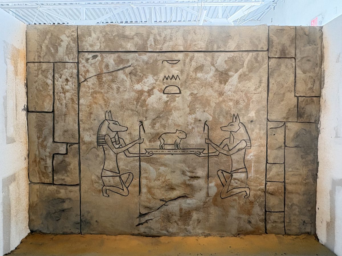 Hand-carved relief scene showing two jackal-headed figures weighing a small capybara on a scale, in an Ancient Egyptian art style